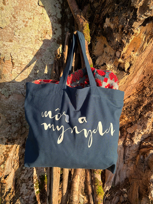 Deep teal tote bag with white text hanging on a tree branch. Welsh Môr a mynydd text Cymraeg Cymru. Recycled cotton canvas
